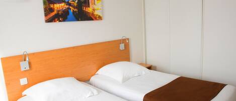 Habitación de hotel con cabecera de madera, dos camas con ropa de cama blanca y marrón, una mesita de noche, un enchufe en la pared y una imagen de una escena de canal.