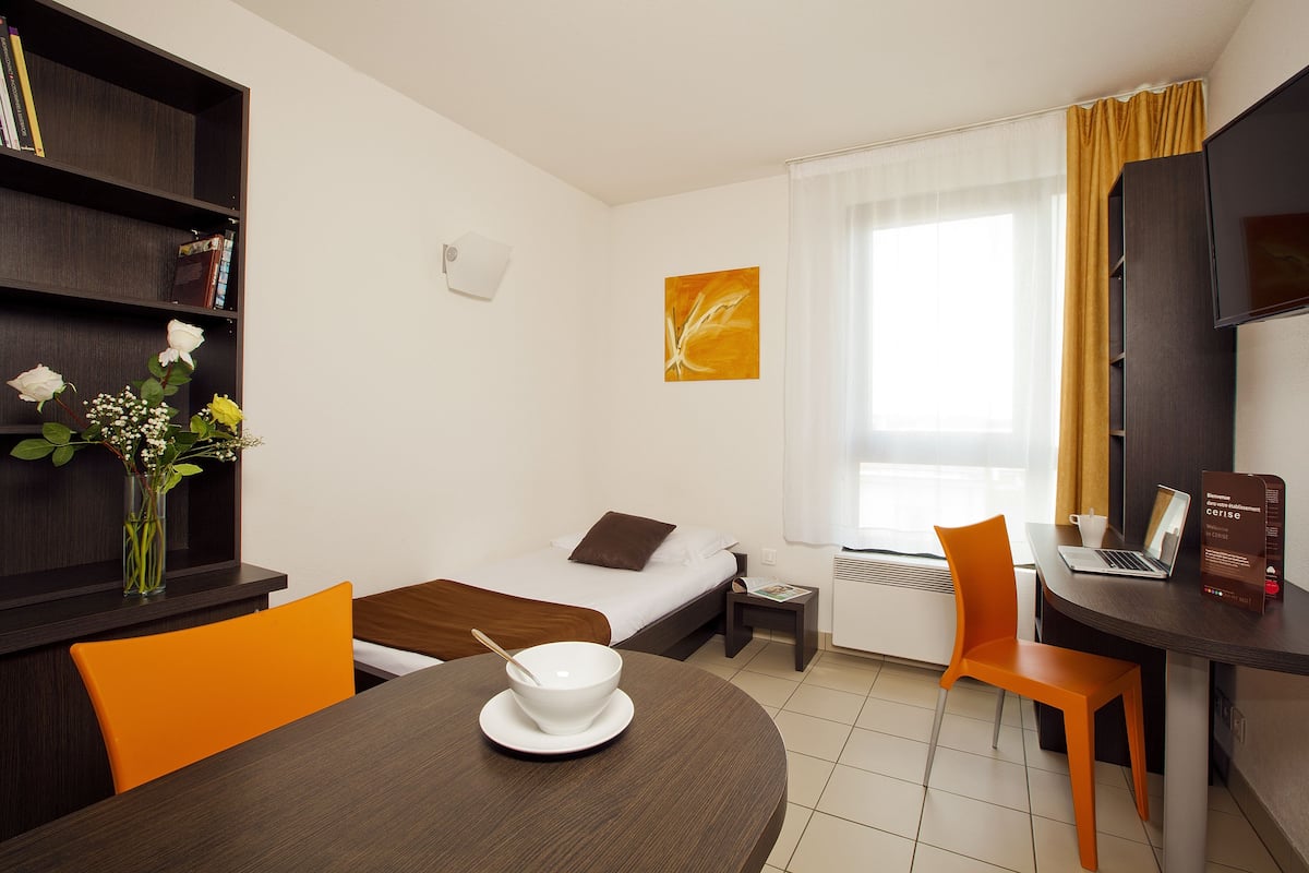 Une chambre d’hôtel comprenant un lit, un bureau avec un ordinateur portable, une table à manger avec un bol, une chaise, une bibliothèque et une fenêtre avec des rideaux.