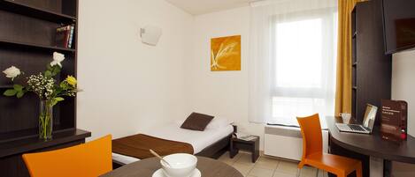 A hotel room with a bed, a desk with a laptop, a dining table with a bowl, a chair, a bookshelf, and a window with curtains.