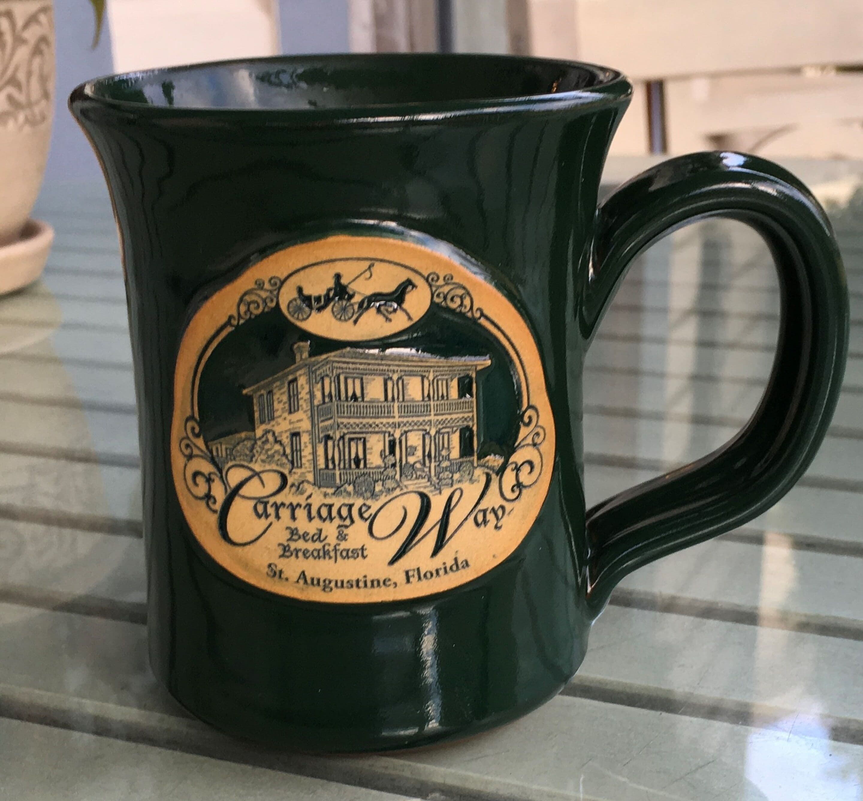 A dark green mug with a vintage design, featuring a horse-drawn carriage and a building illustration, labeled "Carriage Way Bed & Breakfast St. Augustine, Florida."