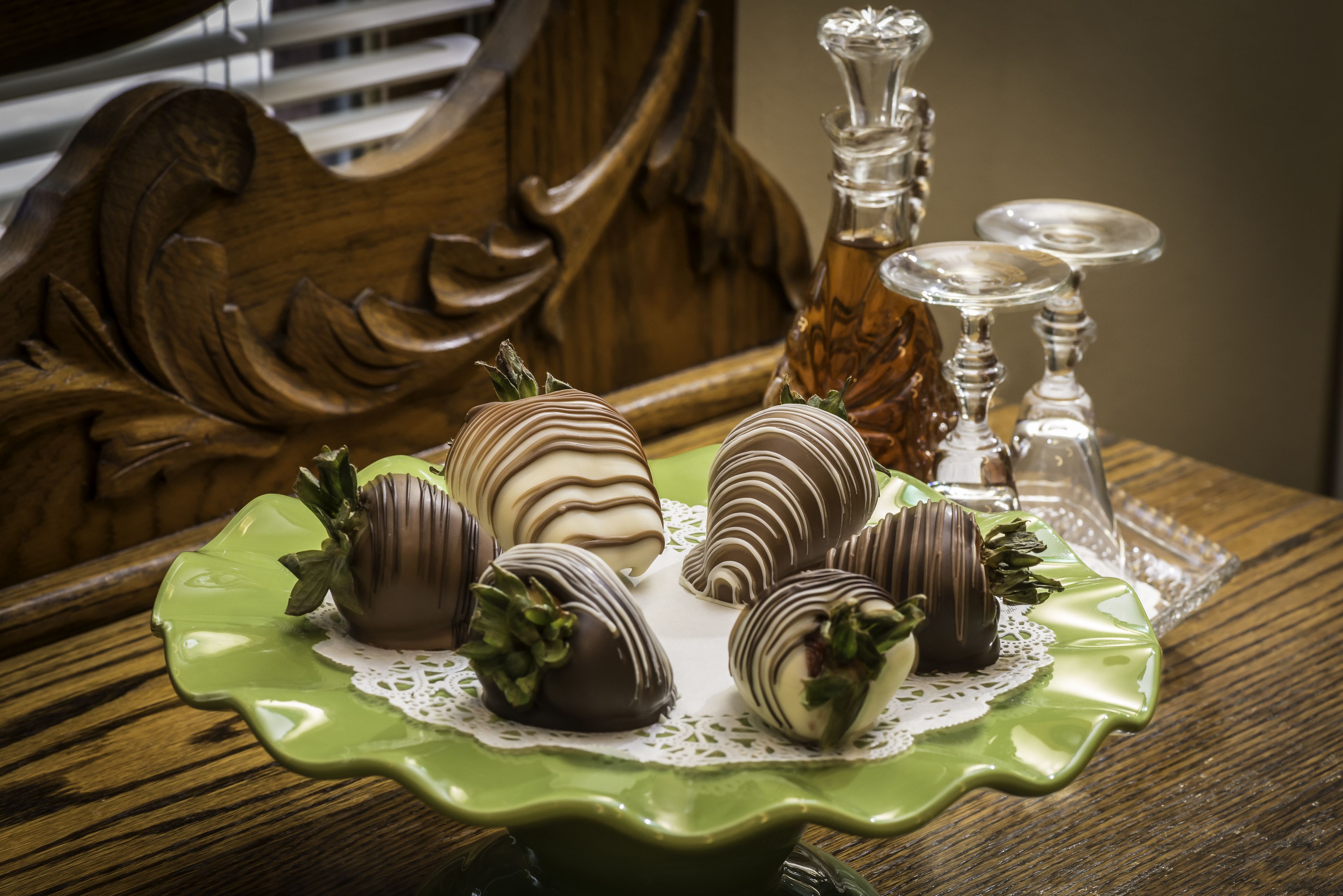 A table with a green plate holding chocolate-covered strawberries, a glass bottle, and two silver candlesticks.