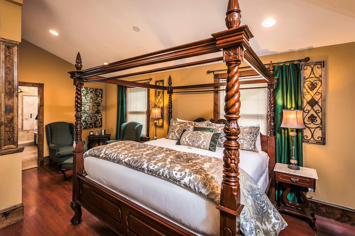 A four-poster bed with a wooden frame, a green upholstered chair, a bedside table with a lamp, and a wall-mounted decorative piece.