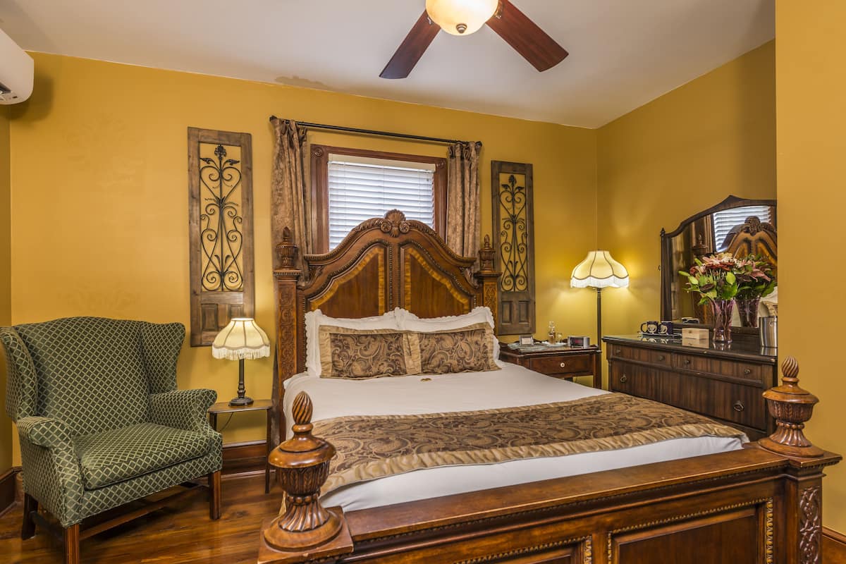 A bedroom with a wooden bed, a green patterned armchair, a nightstand with a lamp, and a dresser with a mirror.