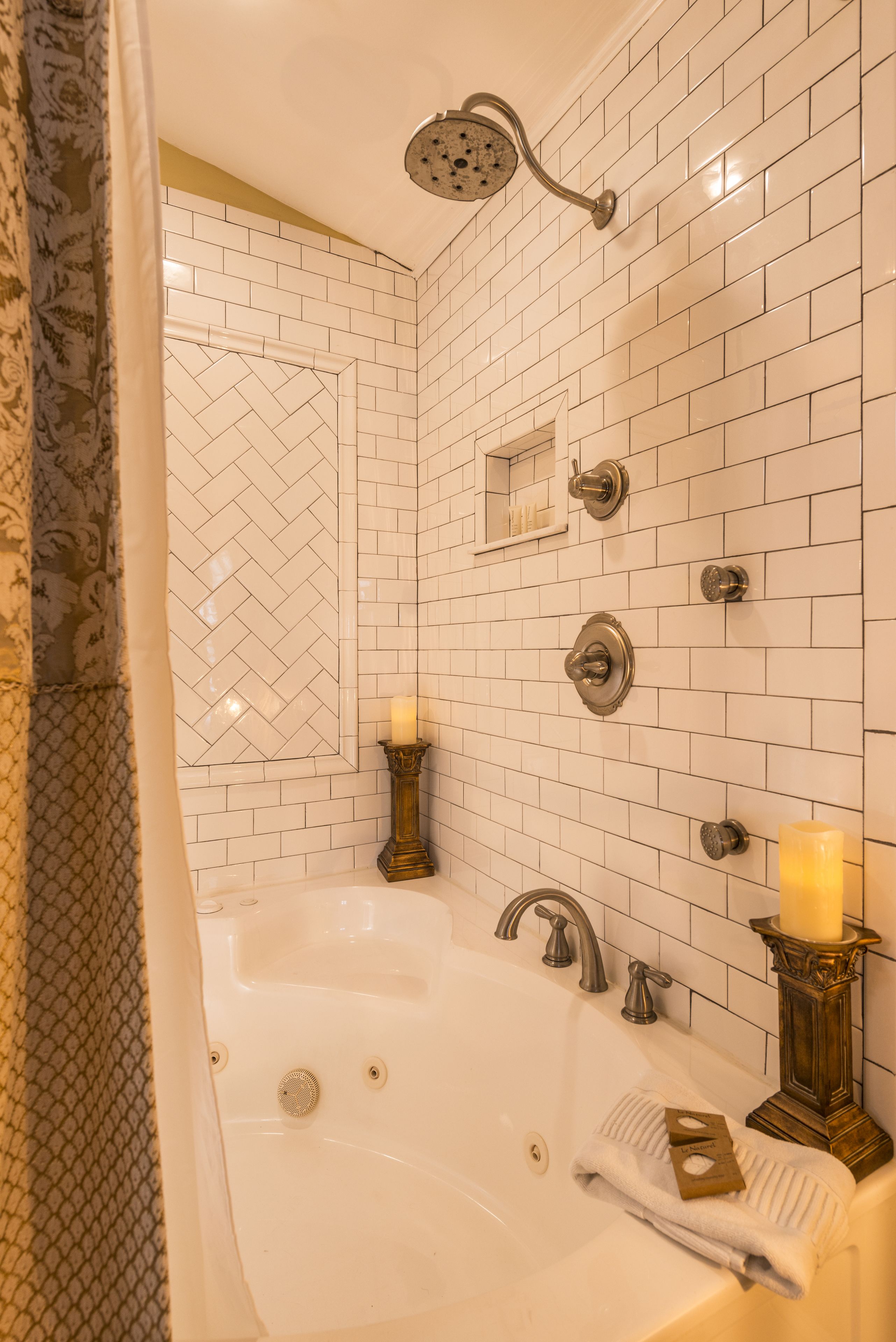 Un baño con un patrón de baldosas blancas en diseño de espiga, una bañera independiente, un cabezal de ducha y dos velas sobre soportes decorativos.