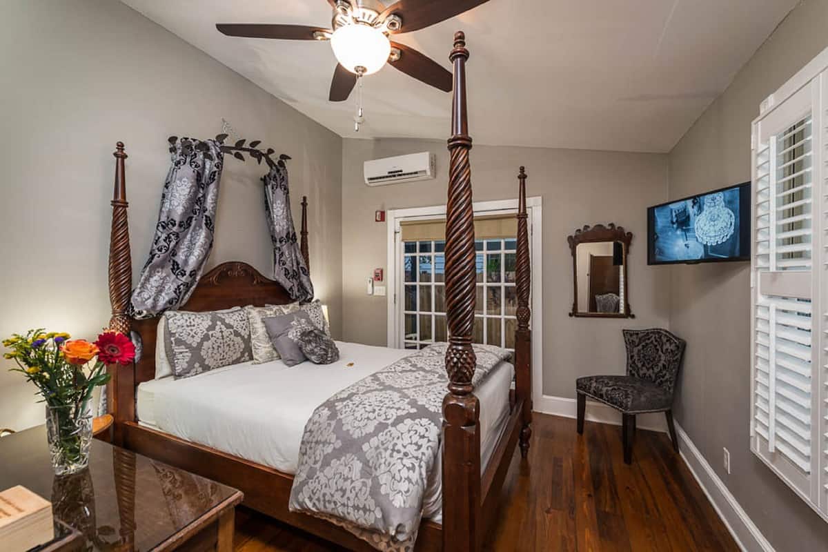 A four-poster bed with a ceiling fan, a television on the wall, and a chair in a bedroom with wooden flooring and a window with blinds.