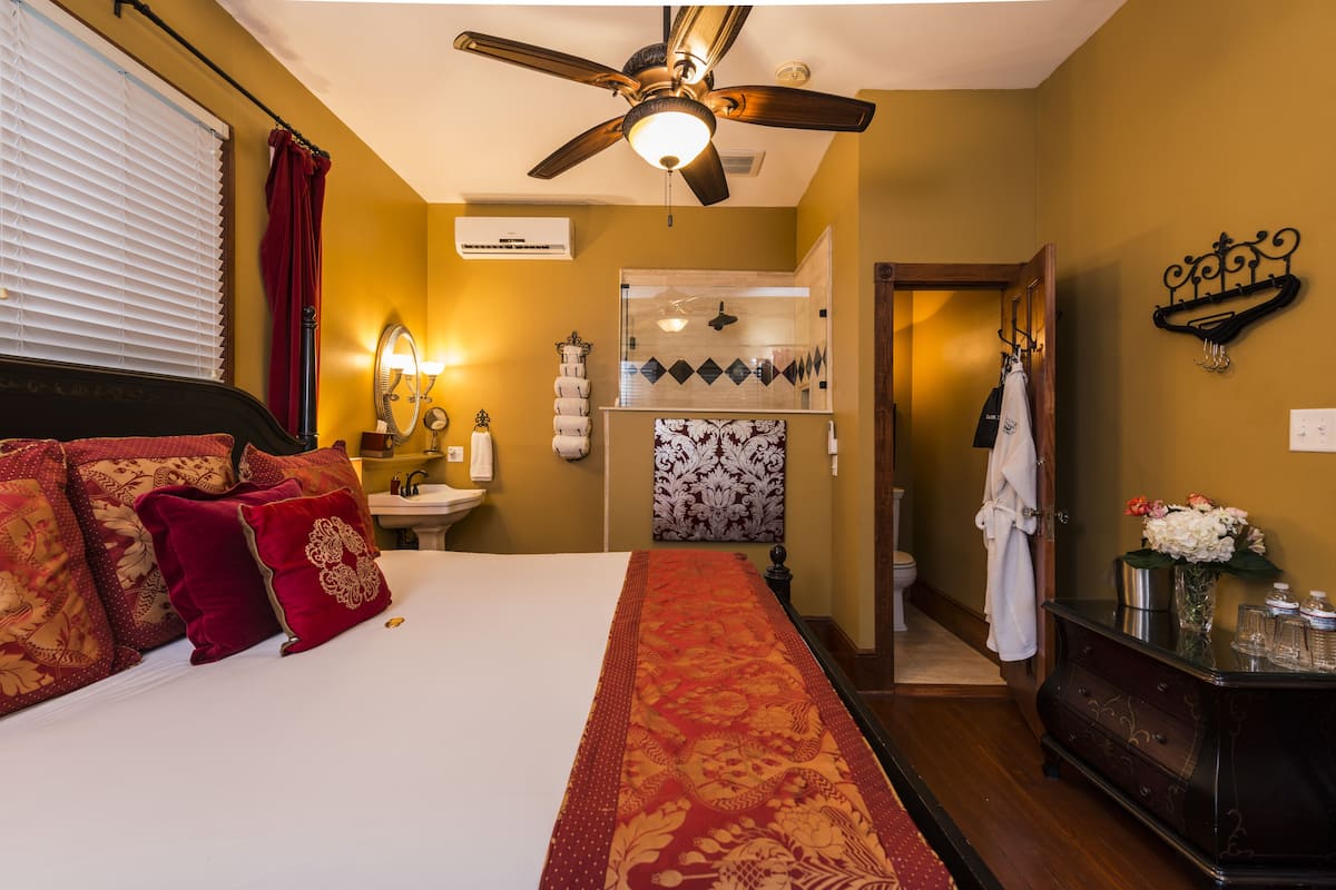 A hotel room with a bed, a ceiling fan, a bathroom with a sink and mirror, and a hallway with a coat rack.