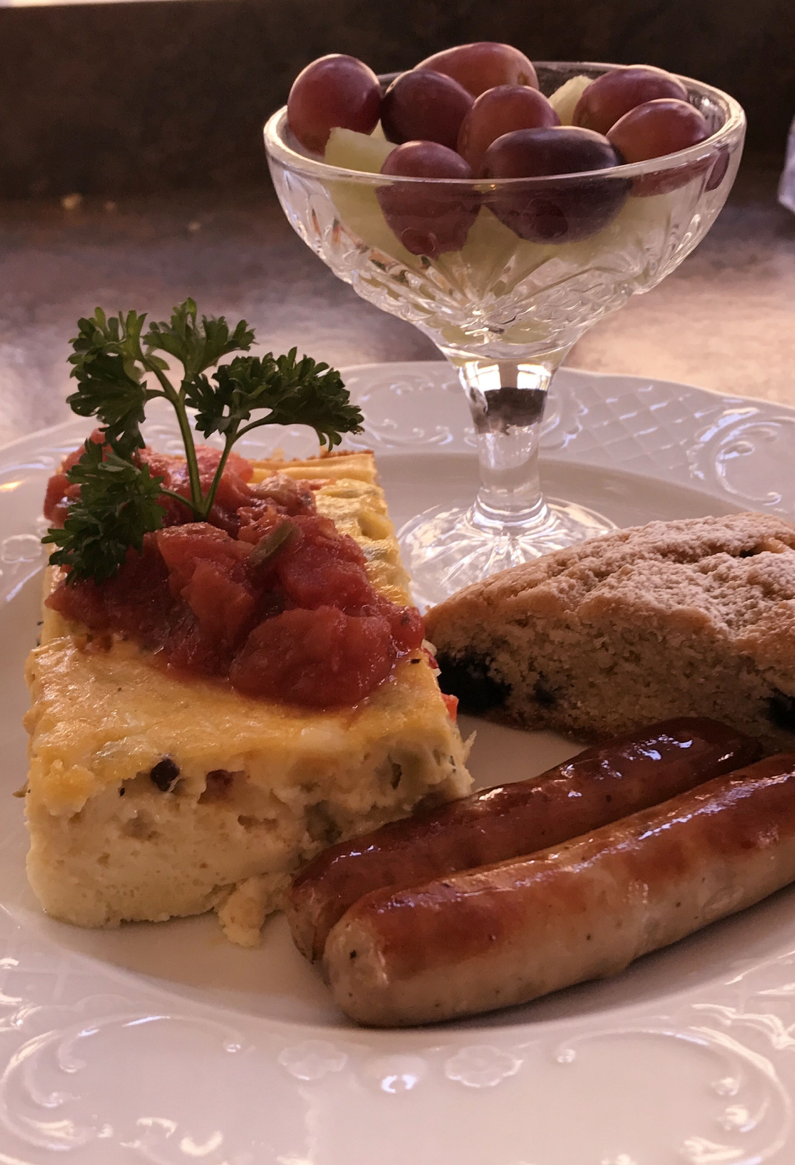 Ein Teller mit einer Scheibe Quiche, Würstchen und einer Schale Trauben.
