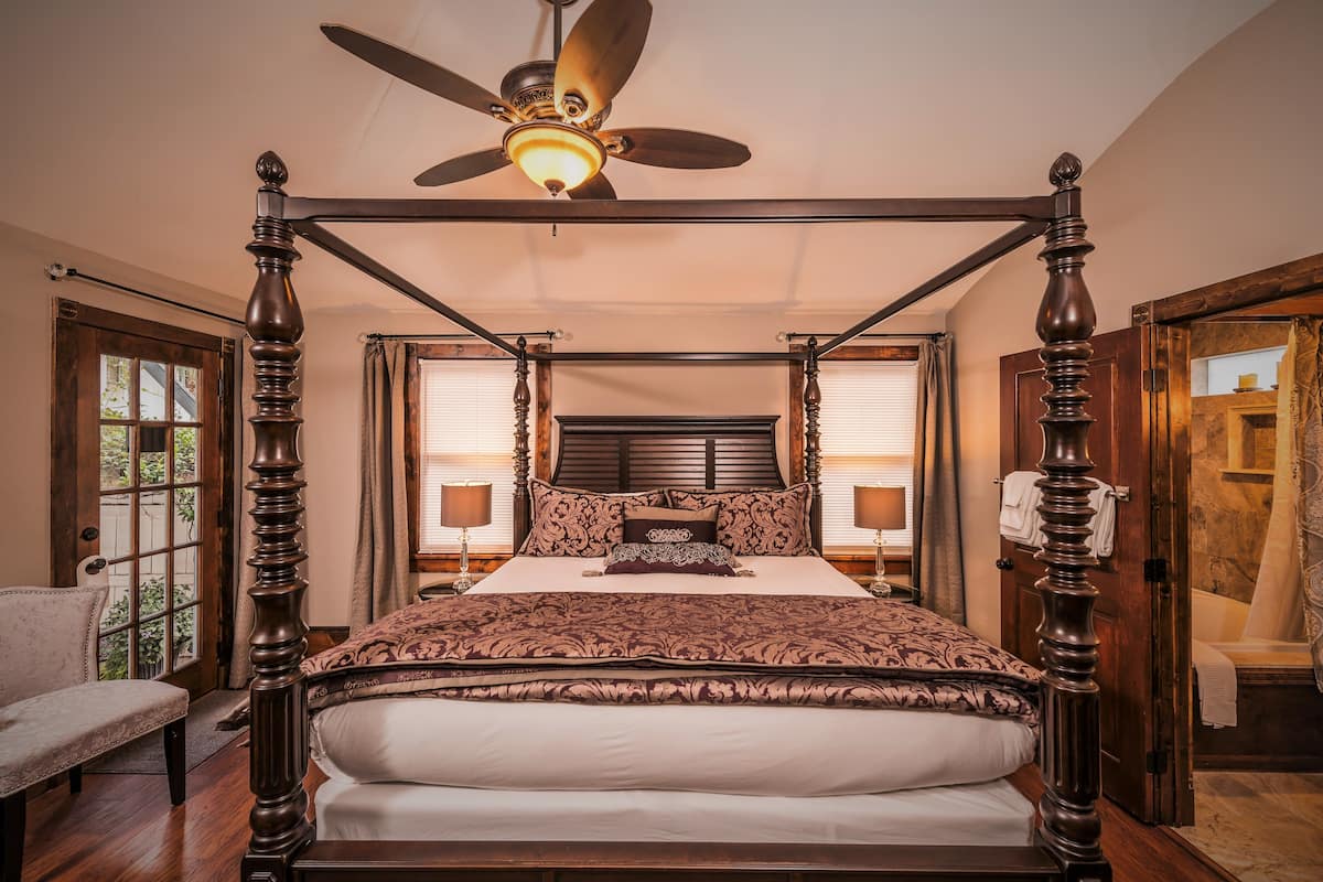 A four-poster bed with a ceiling fan, a wooden floor, and a view of the outdoors through a window.