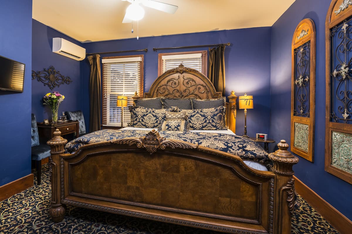 A bedroom with a large bed, a ceiling fan, an air conditioning unit, a bedside table with a lamp, and a wall-mounted TV.
