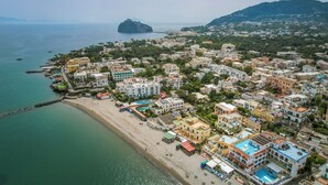 Vista aérea de un pueblo costero con edificios a lo largo de la playa, una colina destacada al fondo y una pequeña isla frente a la costa.