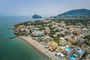 An aerial view of a coastal town with buildings along the beach, a prominent hill in the background, and a small island off the coast.