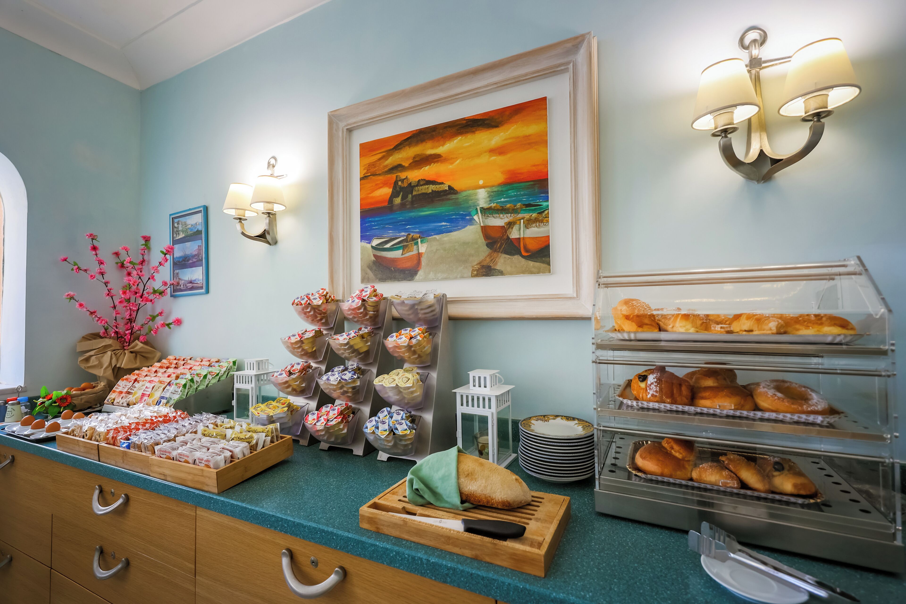 A breakfast buffet with various pastries and bread, a painting of a sunset over the sea, and a wall-mounted light fixture.