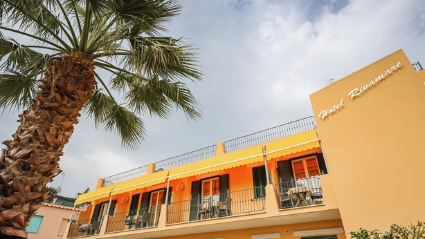 Un hotel con fachada amarilla, balcones con mesas al aire libre y una palmera.