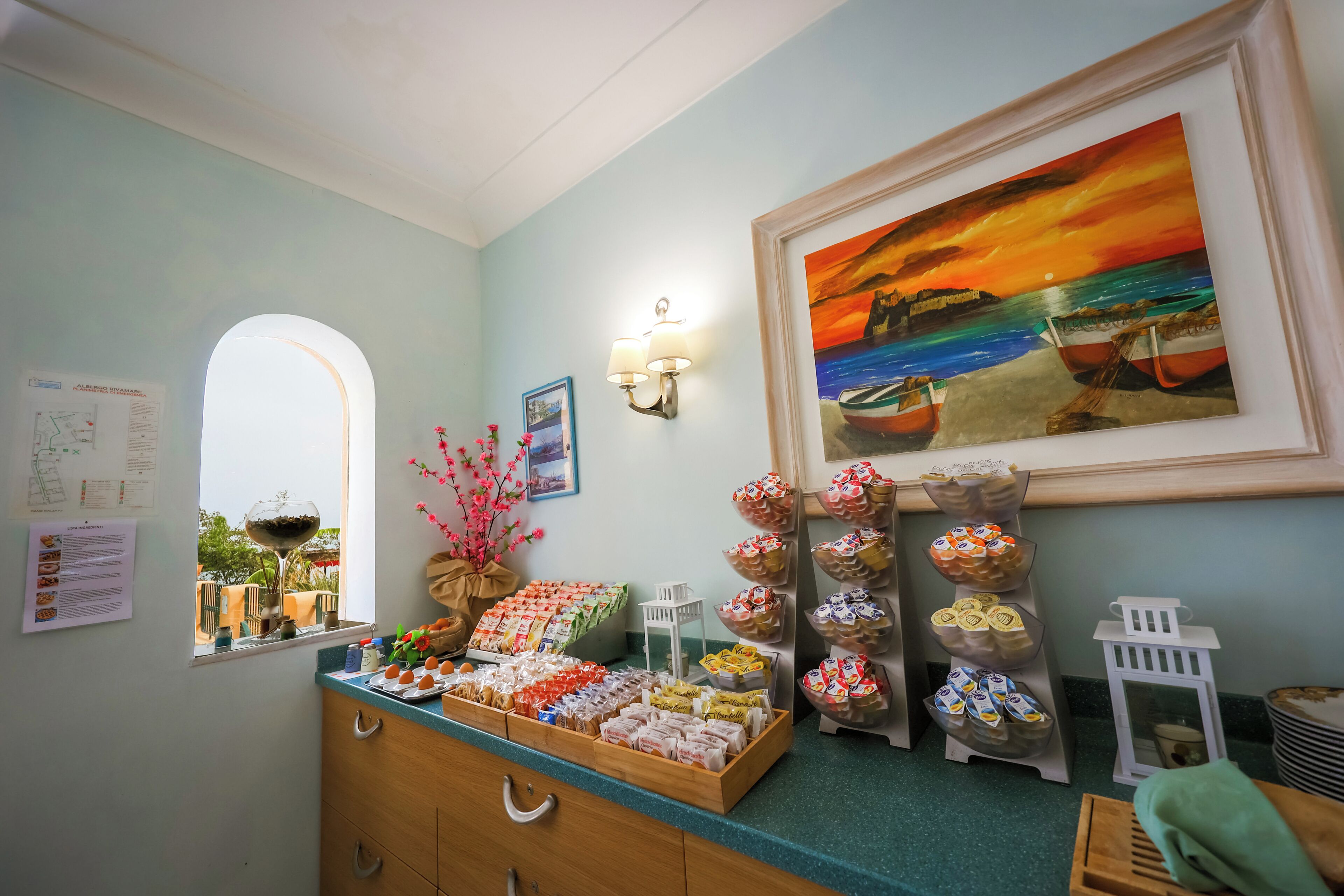A breakfast buffet with various food items, a painting of a sunset over the sea, and a window with a view of the outdoors.