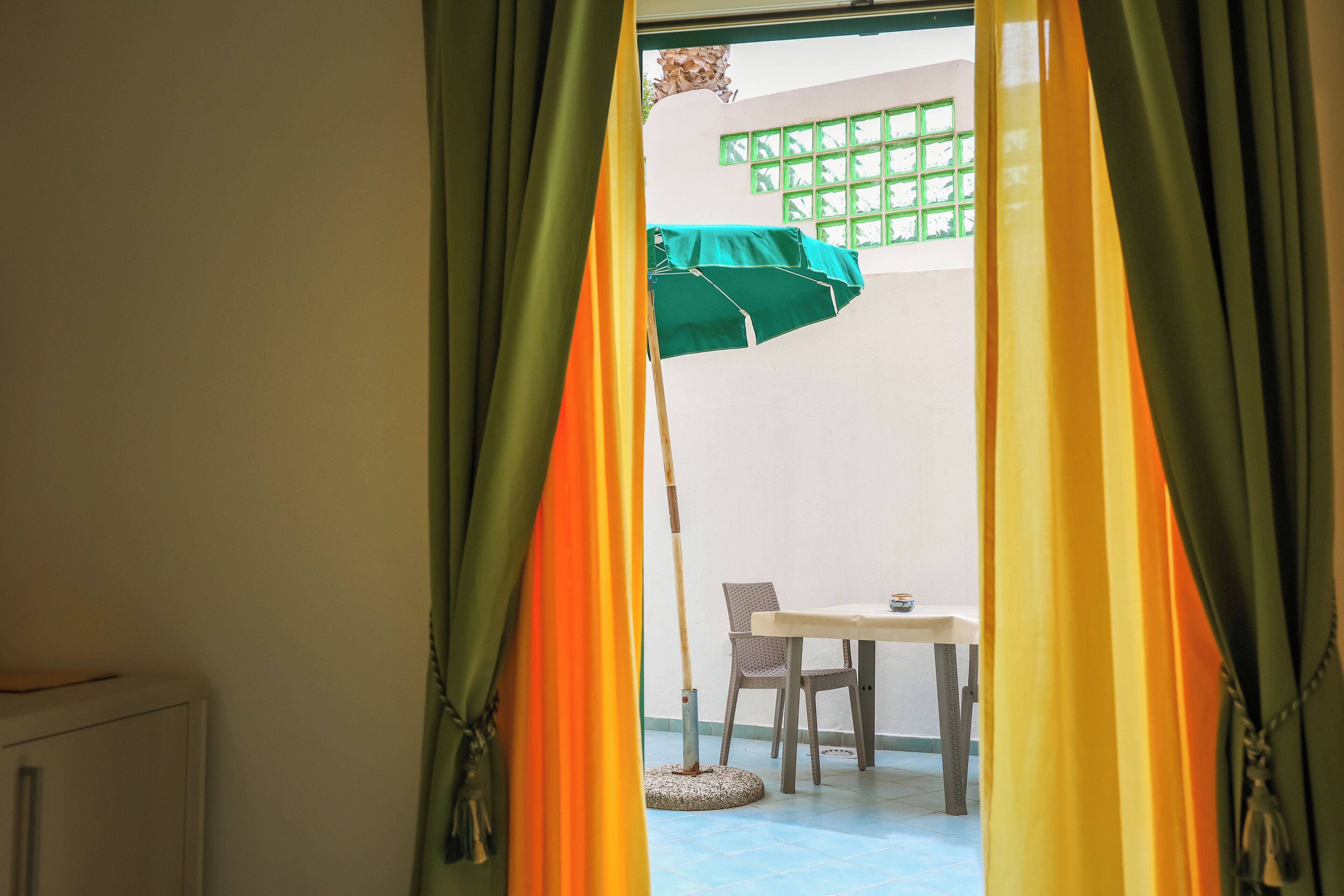 Een kamer met een tafel en stoelen, een raam met een ruitpatroon in het groen, en een deur met groene en gele gordijnen.