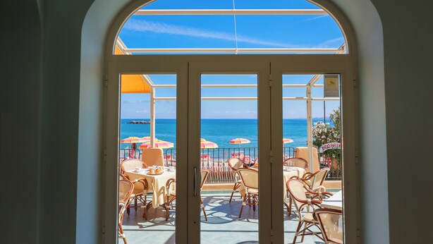 A view through a glass door to an outdoor seating area with tables and chairs, overlooking the sea. - Hotel Rivamare (Ischia)