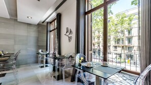Un comedor moderno con sillas y mesas de vidrio transparente, ventanas amplias con vistas a edificios y árboles, y una hoja decorativa en la pared.