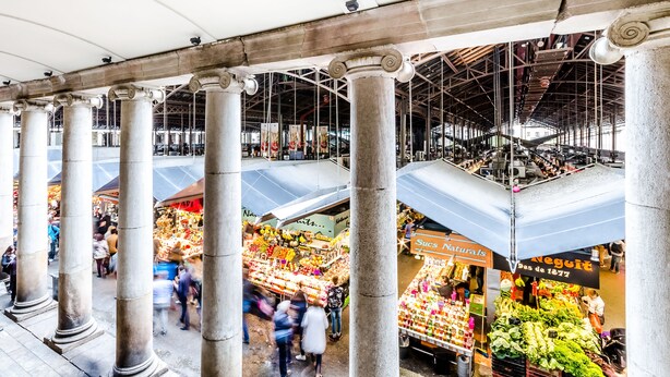 Un mercado interior con varios niveles, con columnas y diferentes puestos que ofrecen frutas y verduras.