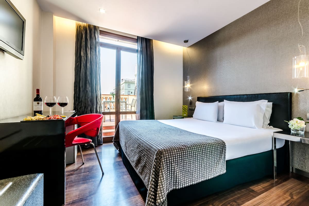 A hotel room with a large bed, a desk with two wine glasses and a bottle, a red chair, and a view of buildings through the window.