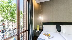 Habitación de hotel con un ventanal grande, cama con ropa blanca, mesita de noche con lámpara y bandeja con artículos de desayuno.