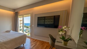 Une chambre d’hôtel comprenant un lit, un bureau avec une chaise, une télévision et une vue sur la mer, visible à travers une grande fenêtre.