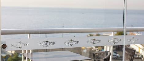 Restaurante con vista al mar, con mesas y sillas, y una barandilla con el rótulo 'ROOF TOP'.