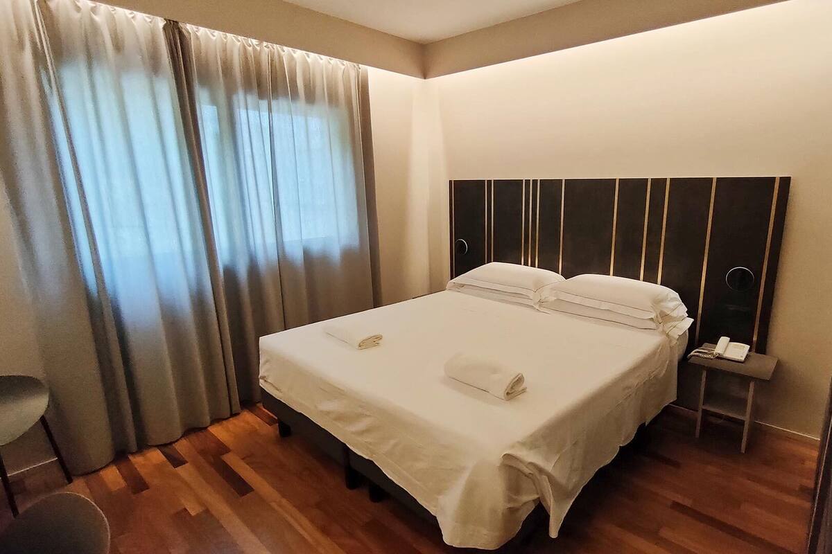 Ein Hotelzimmer mit einem Bett, Vorhängen, einem Nachttisch mit Telefon und einem Sessel.
