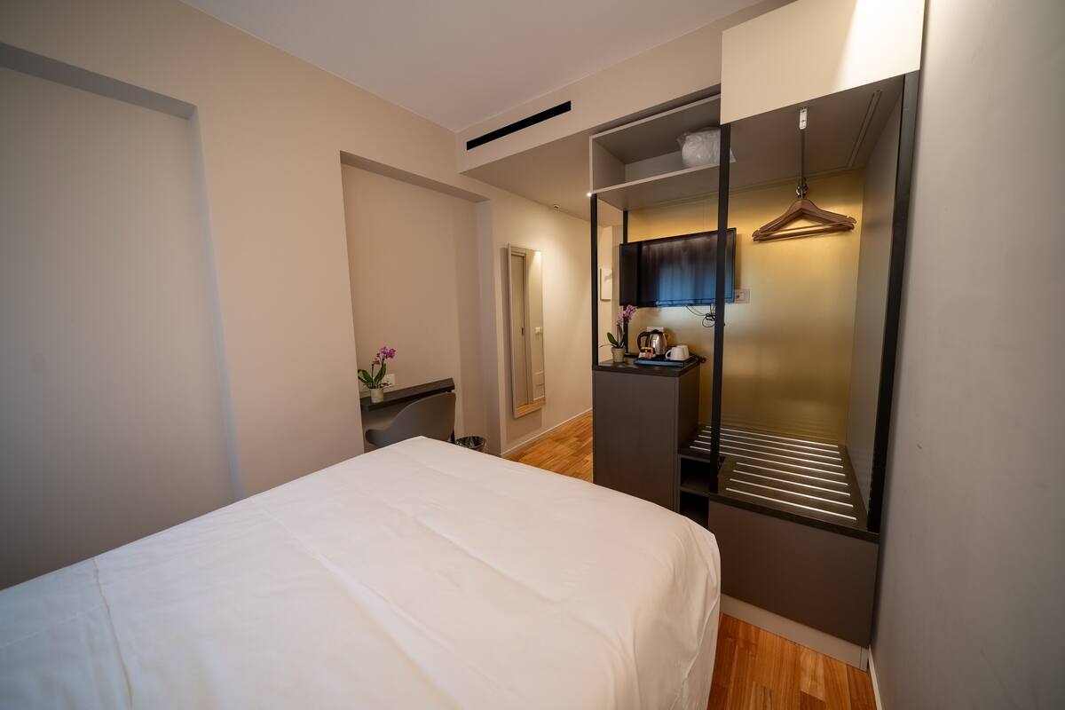 Ein Hotelzimmer mit Bett, Kleiderschrank, Schreibtisch mit Stuhl und einem kleinen Tisch mit Blumenvase.