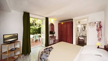 Una habitación de hotel con cama, televisor sobre un soporte de madera, baño con espejo y lavamanos, y vistas al exterior a través de una puerta de cristal.