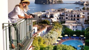 Un balcón con vistas a un pueblo costero, una piscina y una construcción que parece un castillo en un acantilado.