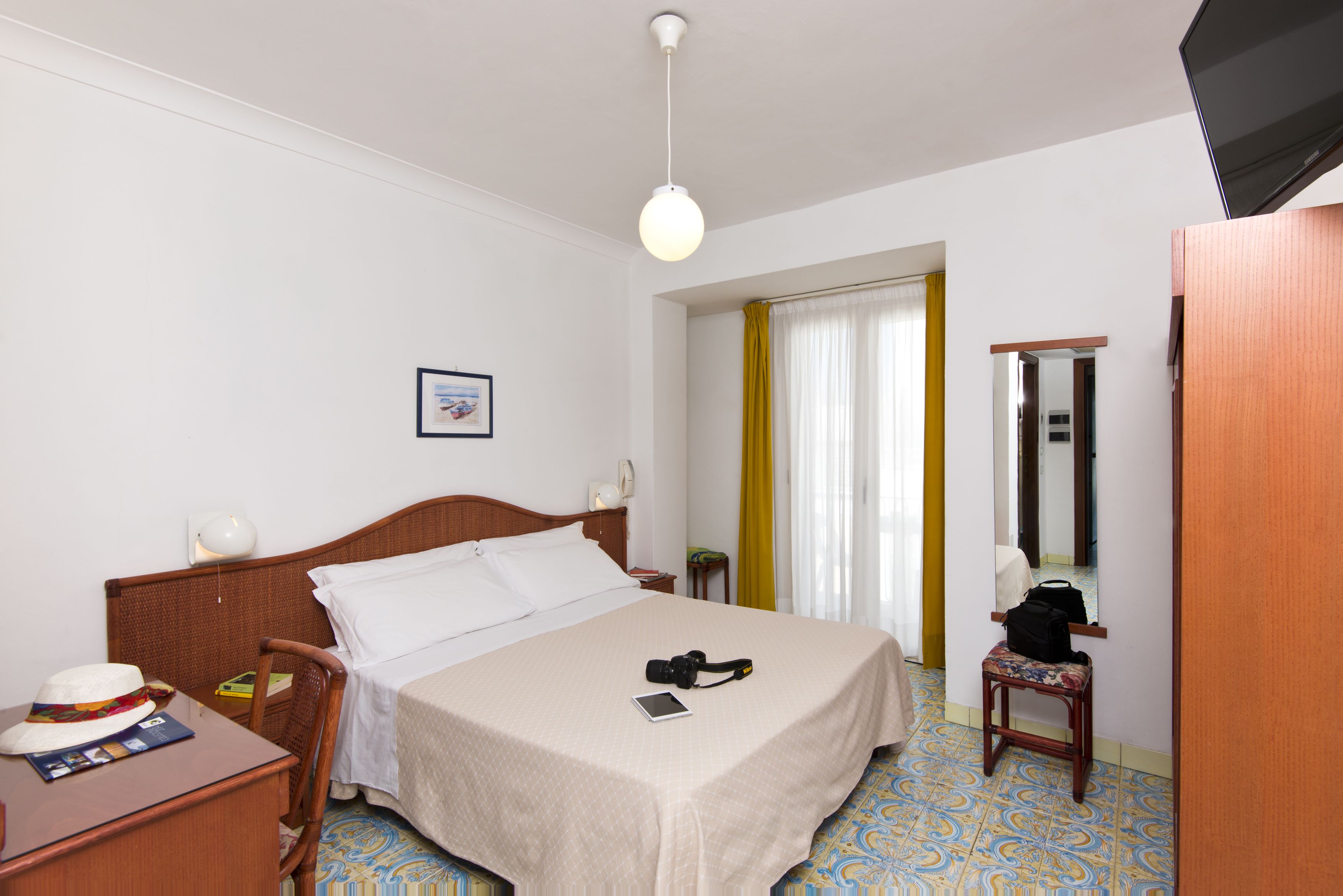 Una camera d'albergo con un letto, una scrivania con un cappello e un libro, una sedia, un tavolino, uno specchio e una finestra con tende.