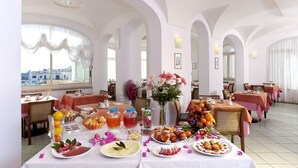 Un comedor con mesas dispuestas para el desayuno, que incluye bollería, frutas y bebidas.