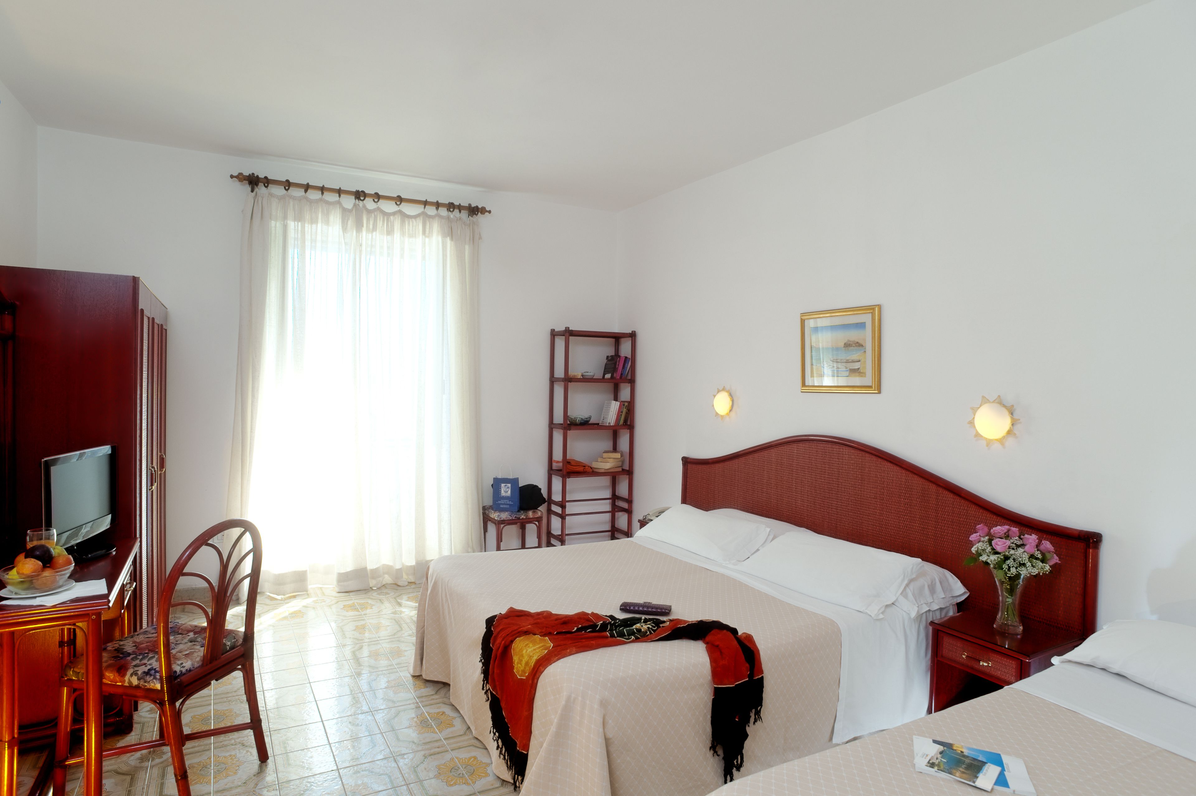 A hotel room with a bed, a desk with a computer, a chair, a bookshelf, and a window with curtains.
