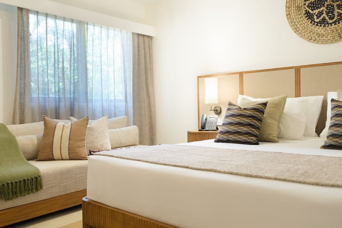 A hotel room with a bed, a sofa, a bedside table with a lamp, and a window with curtains.