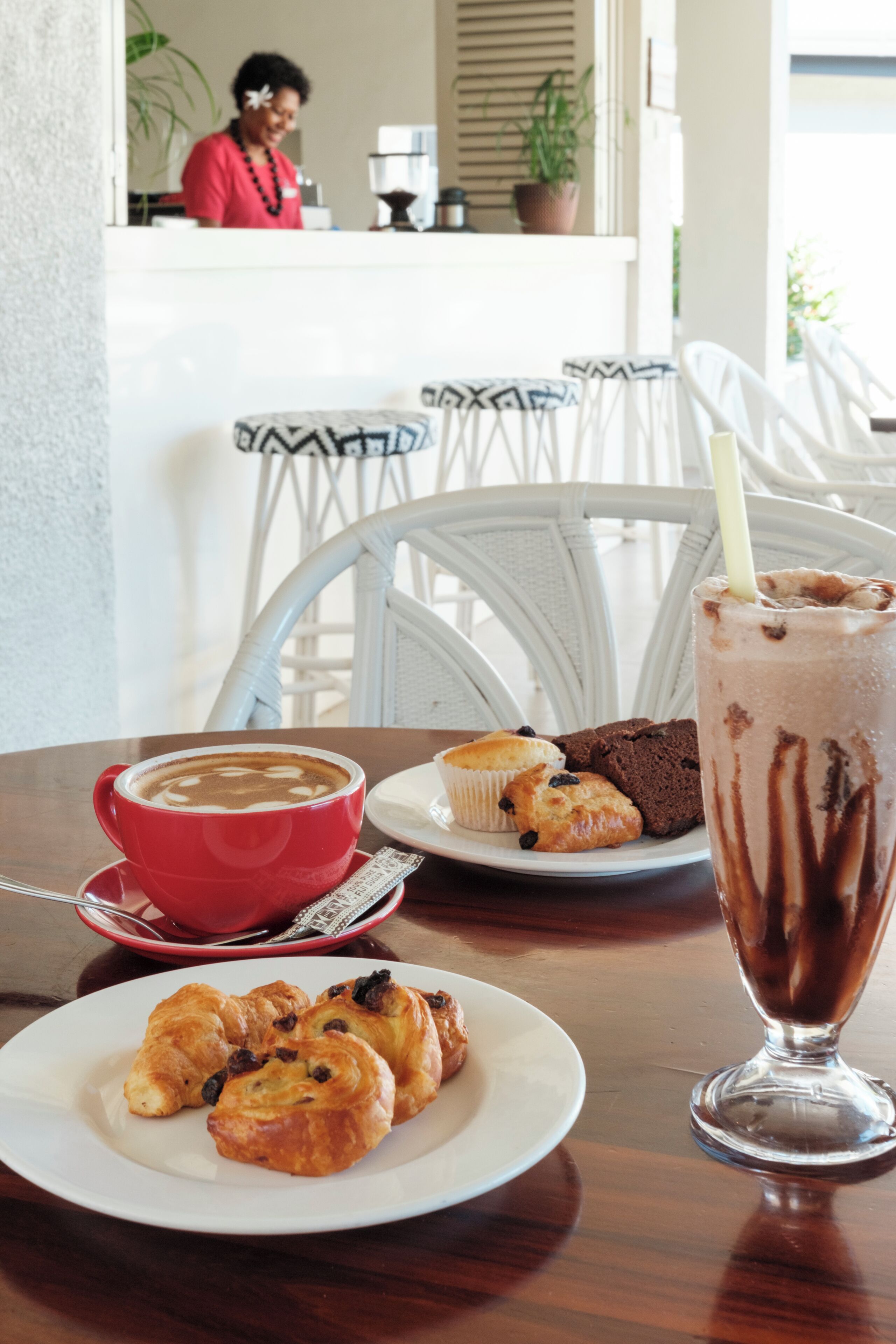 A dining table with a red cup of coffee, a plate of muffins, and a glass of chocolate milkshake.