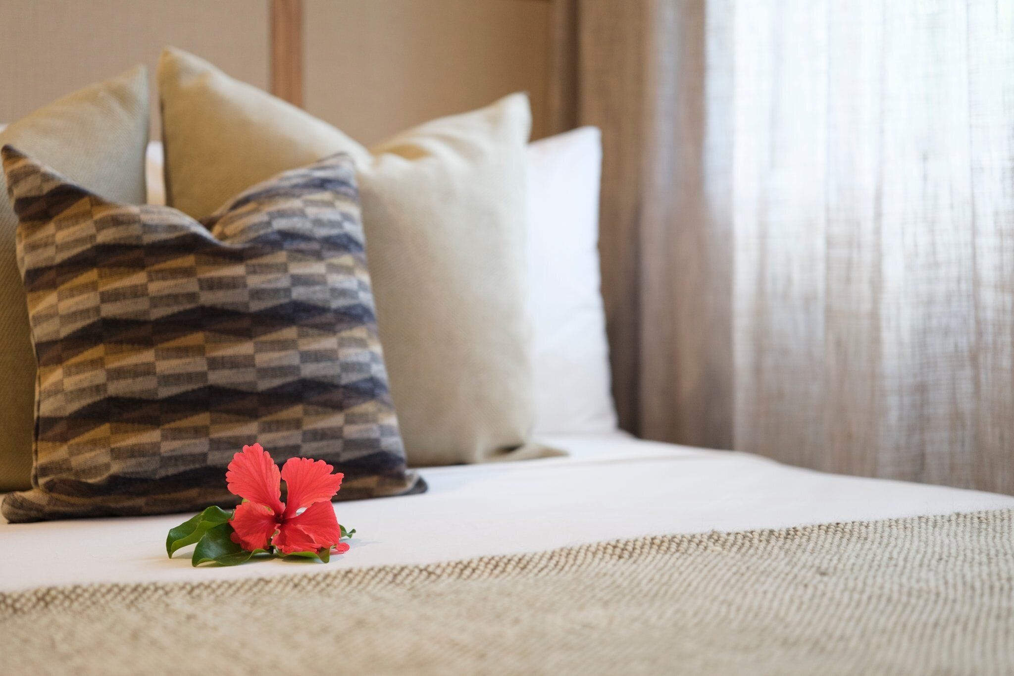 A bed with a checkered pillow, a plain pillow, and a single red flower on the bedspread.