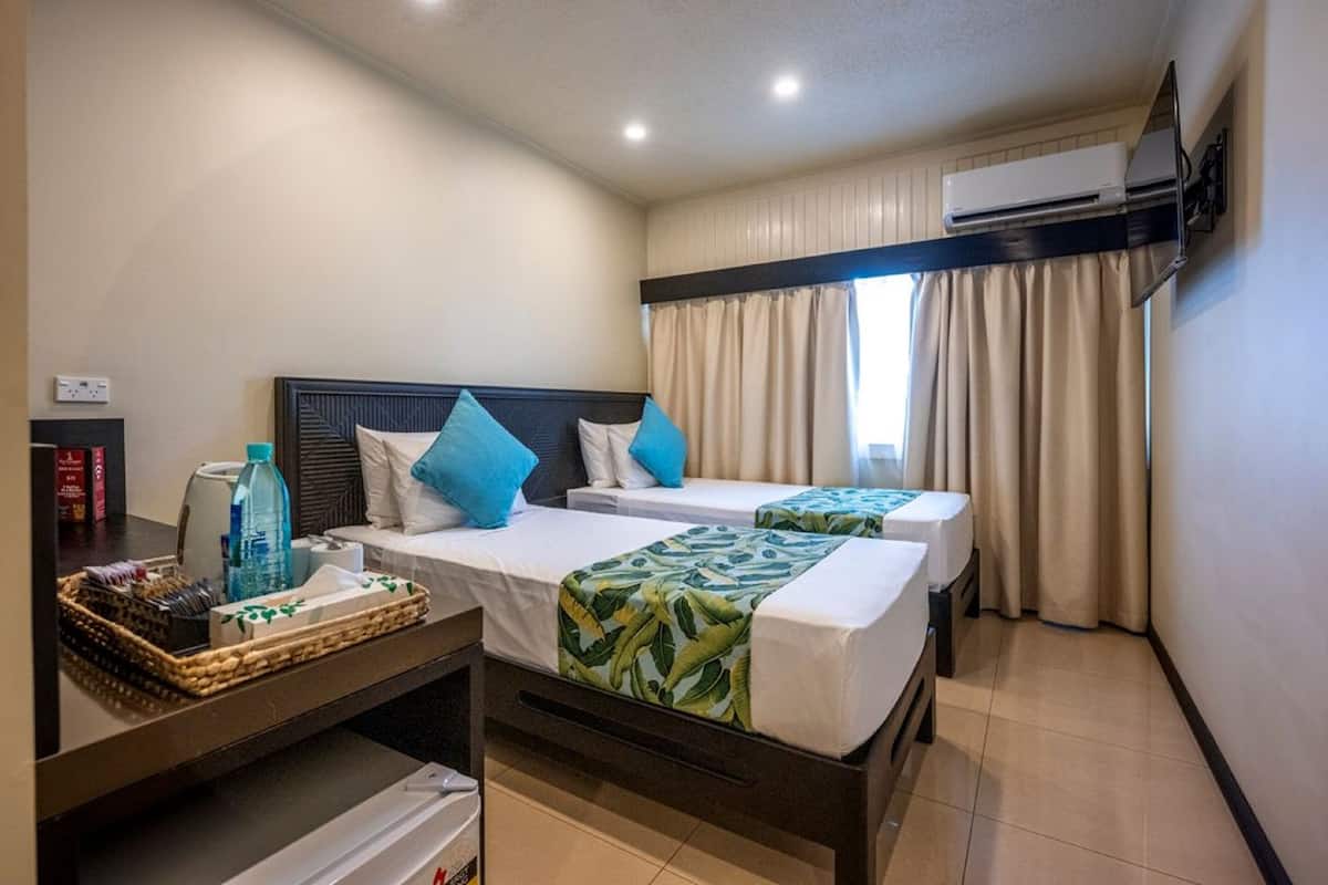 A hotel room with two beds, a small table with a tray of snacks, an air conditioning unit, and a window with curtains.
