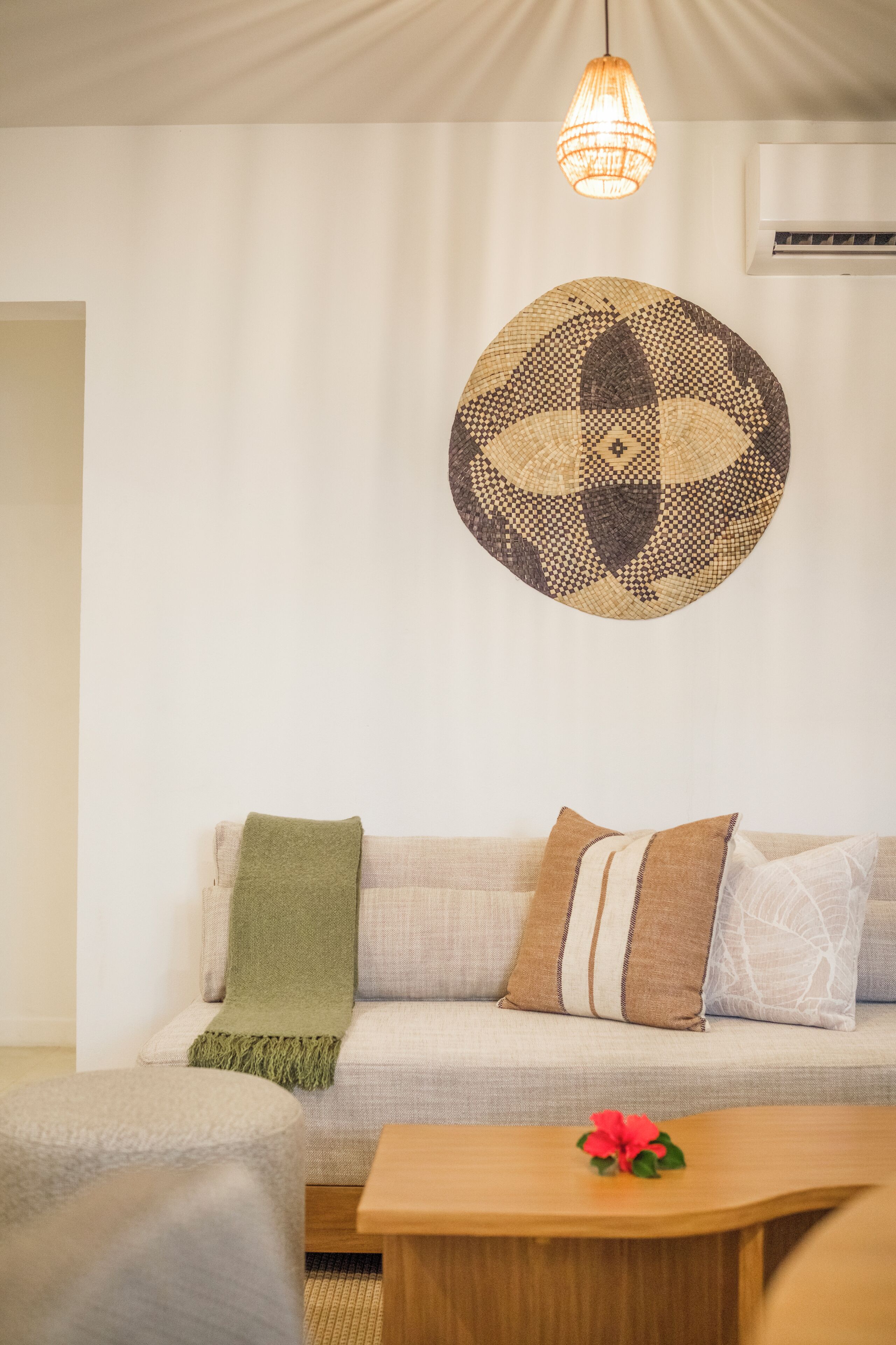 A living room with a sofa, a round woven wall hanging, a wooden coffee table with a flower, and a hanging light fixture.