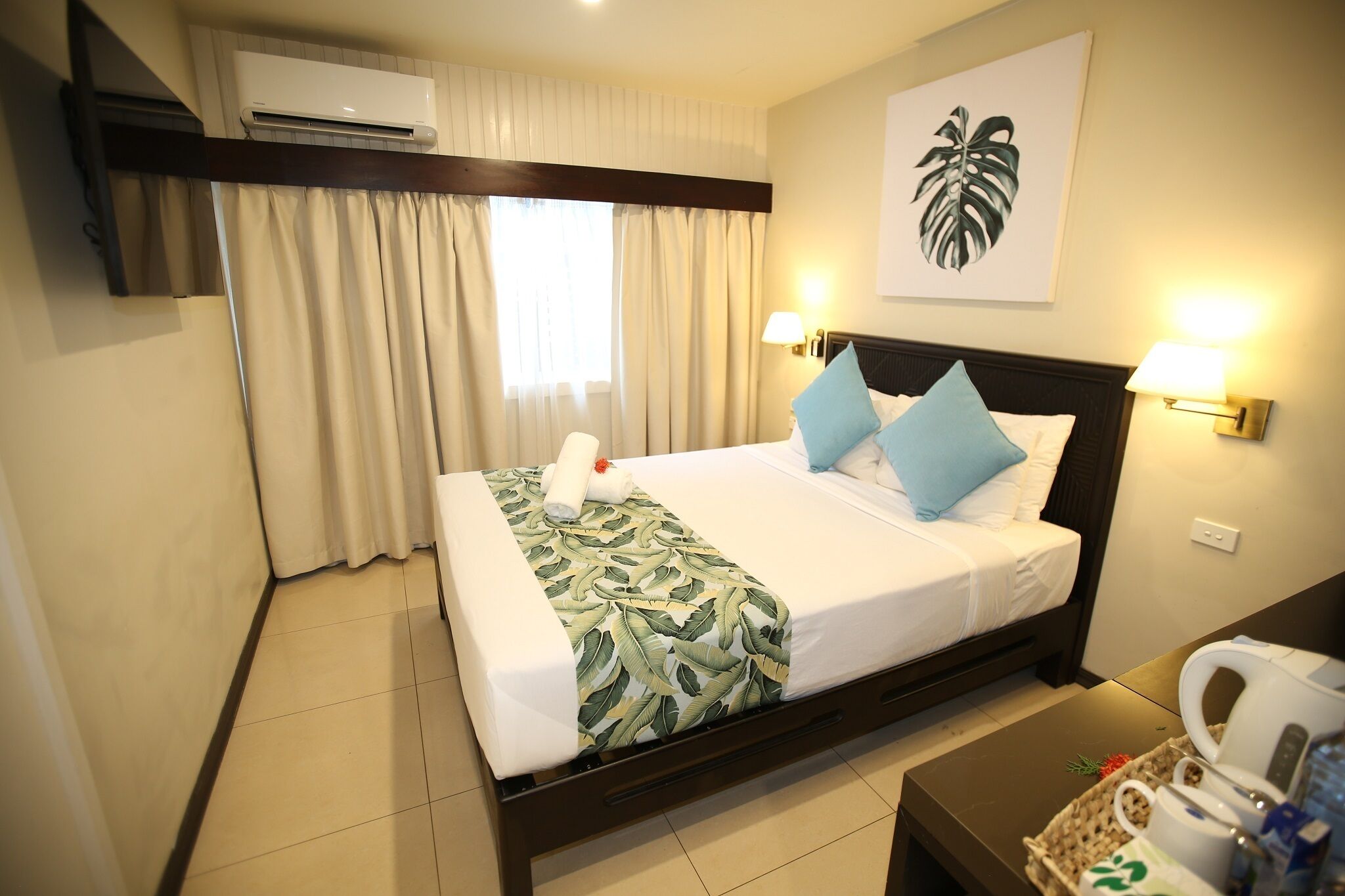 A hotel room with a bed, curtains, an air conditioner, a wall-mounted lamp, and a framed plant.