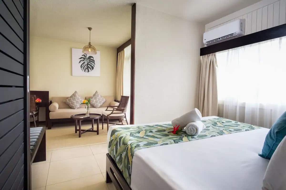 A hotel room with a bed, a sofa, a small table, and a chair. There is an air conditioning unit on the wall and a window with curtains.