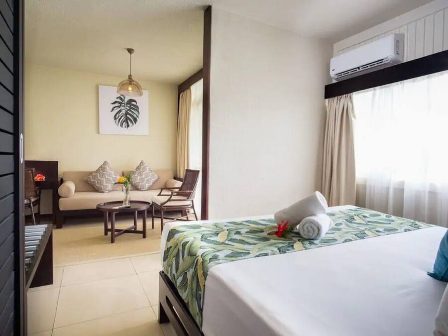 Une chambre d’hôtel avec un lit, un canapé, une petite table et une chaise. Il y a un climatiseur fixé au mur et une fenêtre avec des rideaux.