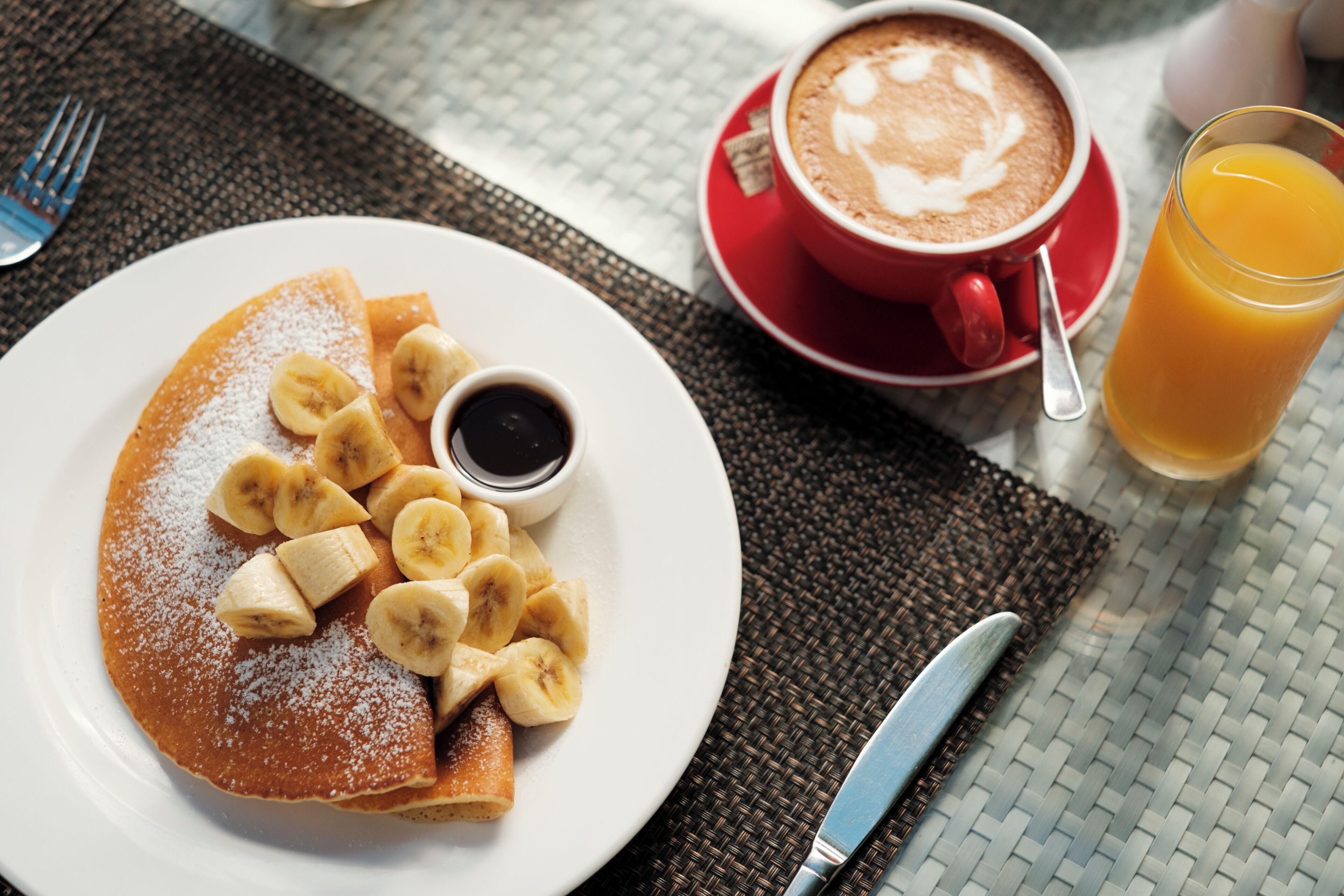 A breakfast setting with a plate of pancakes, bananas, and syrup, a cup of coffee, and a glass of orange juice.