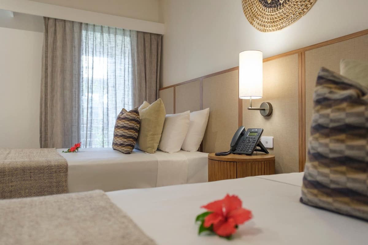 A hotel room with two beds, a bedside table with a phone, a lamp, and a window with curtains.