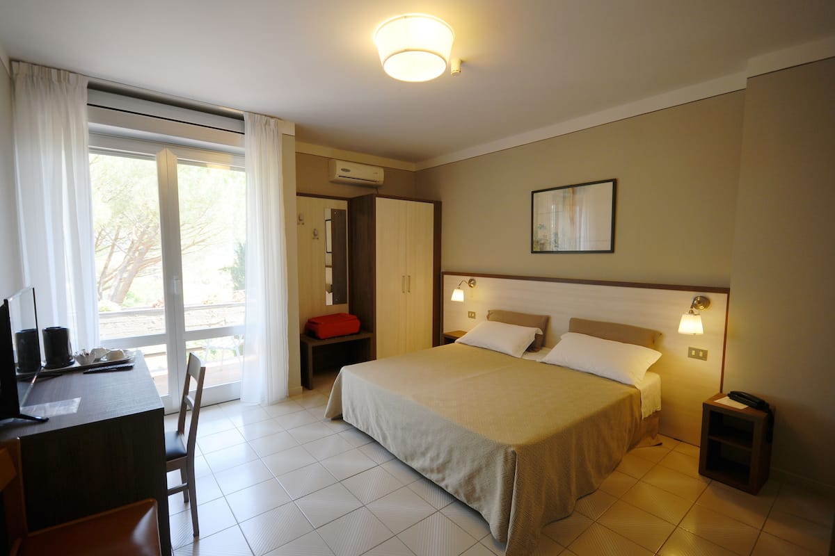 A hotel room with a bed, bedside table, desk, chair, and a view through a window.