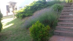 Um jardim com uma escadaria de pedra, um pequeno edifício com cúpula e diversas plantas, incluindo lavanda e flores vermelhas.