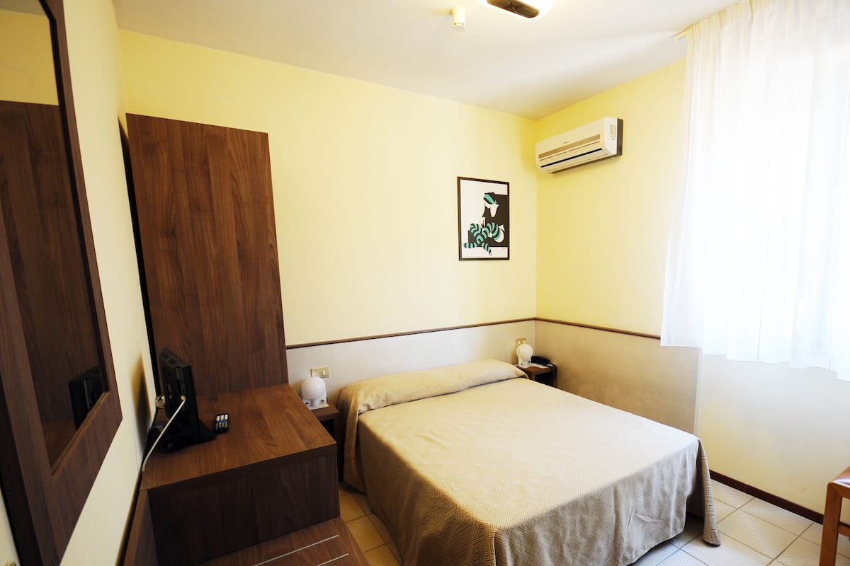 A hotel room with a bed, a desk, a wardrobe, an air conditioner, and a window with curtains.
