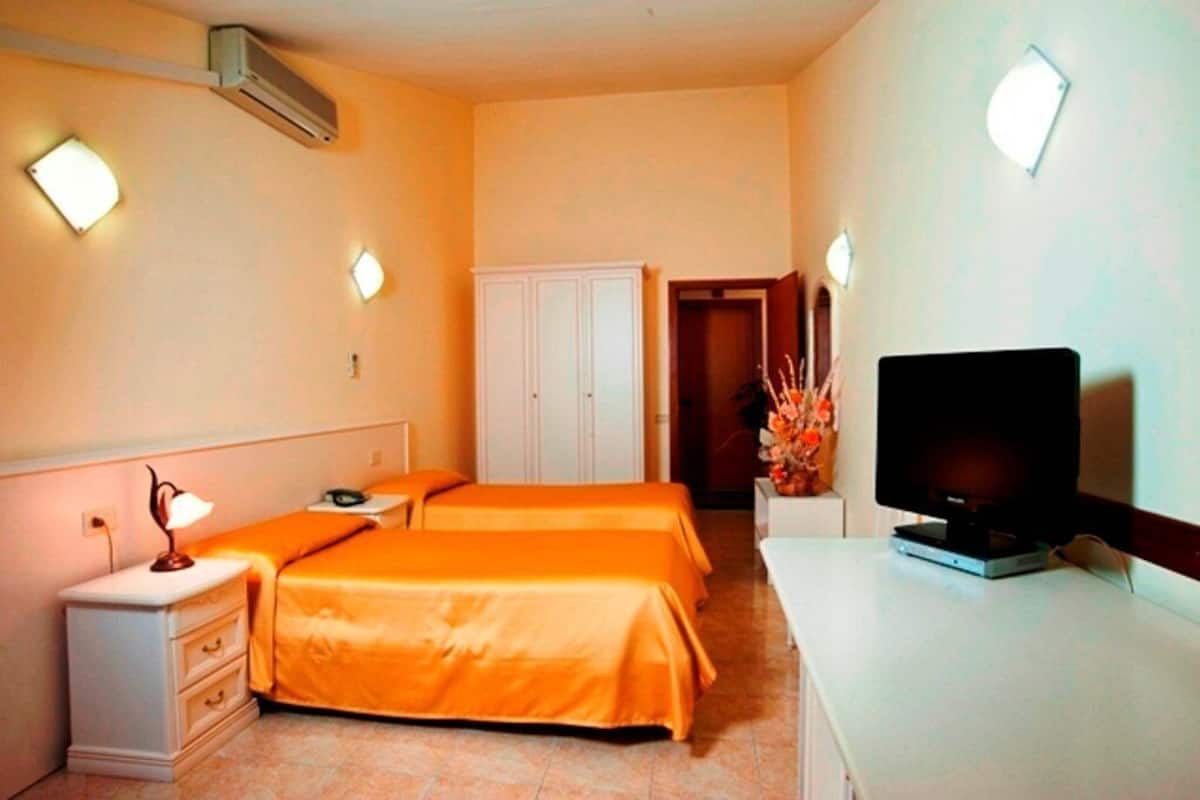 A hotel room with two beds, a desk with a television, a wardrobe, and wall-mounted lights.