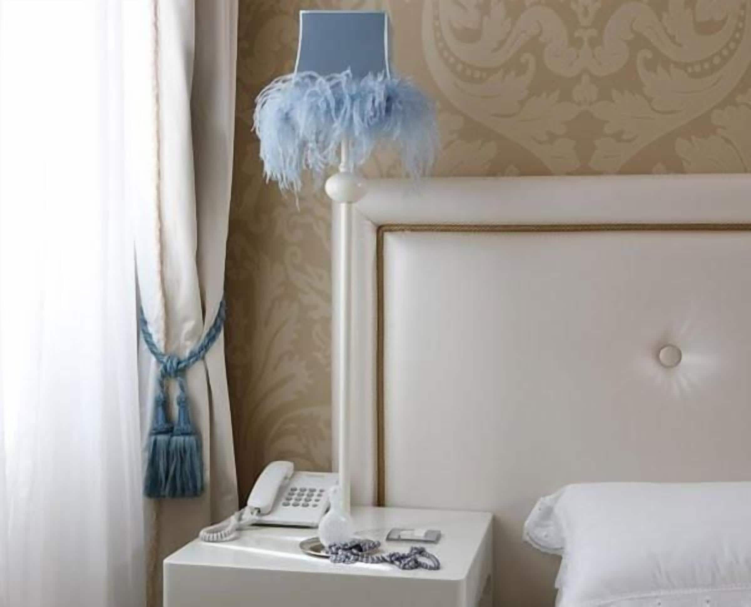 Una habitación de hotel con una cama blanca, una mesita de noche con teléfono y lámpara, y una cortina con flecos azules.