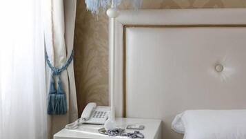 Una habitación de hotel con una cama blanca, una mesita de noche con teléfono y lámpara, y una cortina con flecos azules.