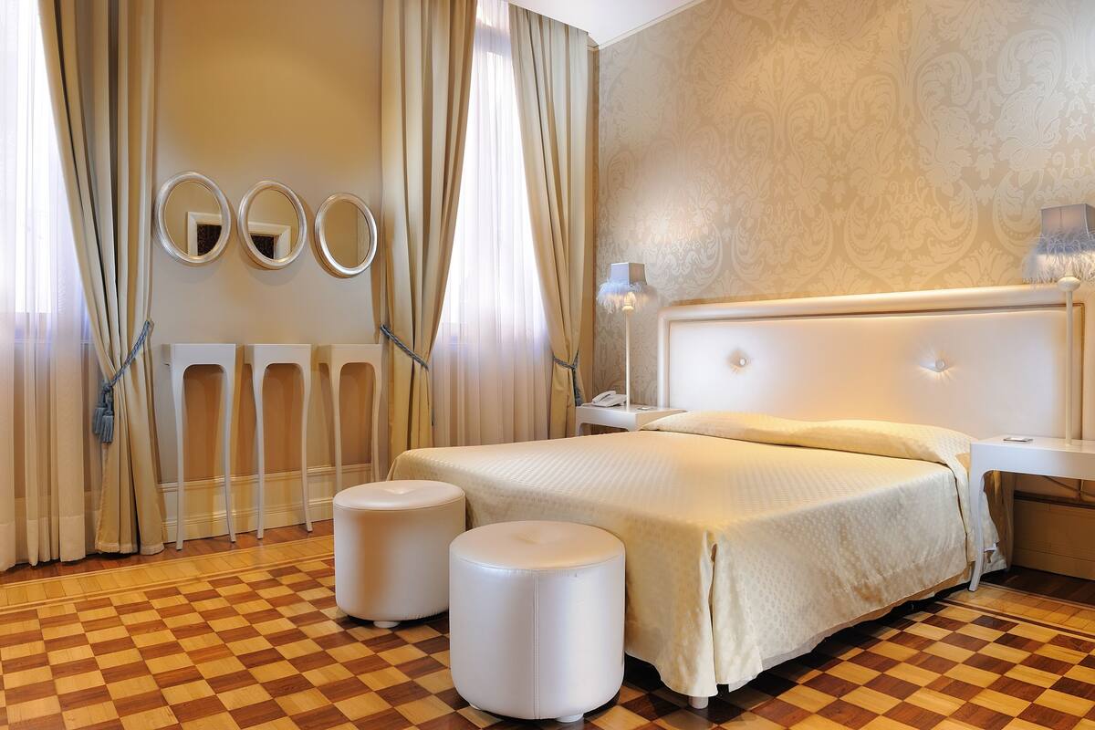Quarto de hotel com chão quadriculado, uma cama grande com cabeceira branca, dois pufes brancos e três espelhos redondos na parede.